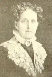 An older white woman wearing a large portrait brooch at her throat, a high lace collar and a lace shawl.