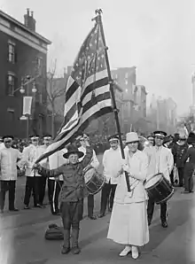 News photograph of a suffrage parade, with a white woman wearing white, holding a large American flag; a boy is helping to hold the flag. Drummers stand behind the two figures in the foreground.