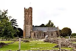 Stone building with square tower. In the foreground are low walls of the ruins amongst the grass.