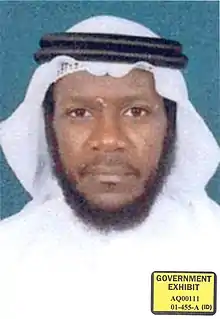 Mustafa al-Hawsawi sitting crosslegged on a green prayer rug. He is wearing a brown pakol hat and a white T-shirt. He is looking directly into the camera.