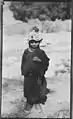 Rumbur Valley, Chitral, NWFP, Pakistan Kalash girl in Rumbur. The girl is dressed in a dark gown and carries jewellery: a ring, a necklace and a headdress (kupa) covered with shells (kauri or cowri). On top of the headdress there is a tassle (probably red) and a second one, smaller, is visible above her forehead. Her shoes are also clearly visible. 1929 approx.