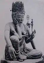 Three-quarter view of a seated statue with six arms. The left leg is bent, touching the ground along its whole length. The right leg is also bent, but with the knee up. The right foot is above the left foot. One of the right hands is touching the right ear, another is lifting a jewel-shaped object in front of the breast. One of the left hands carries a flower, another a small wheel-shaped object.