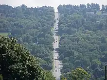 A two-lane highway descends sharply into a valley, where it intersects another roadway at the base of the valley. From there, it ascends the opposite side of the valley and disappears into the background.