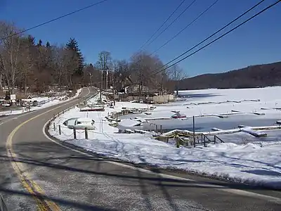 A road straddling the bank of a frozen lake. The surrounding ground is covered by thin layer of snow. Powerlines are visible overhead