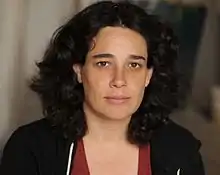 Headshot of Levari; she is looking into the camera, and has shoulder-length dark hair. She is wearing a dark jacket over a red scoop-neck top.