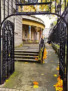 The photo shows the open black gate with the word "Library" in gold letters in the foreground with some yellow leaves that have fallen to the ground. The half-circle shaped part of the front of the main building of the National Library of Ireland can be seen through the open doorway of the gate.