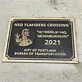 A photo of the Ned Flanders Crossing plaque, with a cartoon of Ned Flanders and the caption HI-DIDDLY-HO, NEIGHBORINOS!"