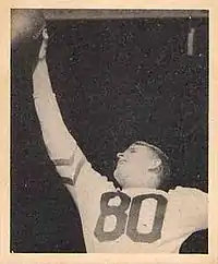 Black and white photograph of Armstong in a light-colored number 80 jersey reaching up with his right hand to catch a football