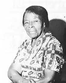 1/2 portrait photograph of a black woman in a patterned, short-sleeved dress whose arms are crossed in her lap.