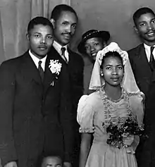 A black and white photograph of a black man and a black woman, standing side by side. He is wearing a suit, she is wearing a white dress.