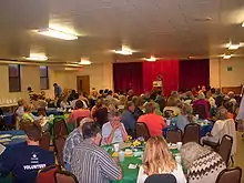 Tables of eight or so people watch a speaker standing before red curtains.