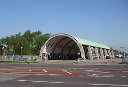 A grey, cylindrical structure with a green roof and a large number of people walking through it with trees in the background all under a blue sky