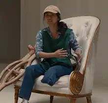 A Vietnamese woman wearing a cap with her left leg missing below the knee sits in a chair with wooden crutches next to her.