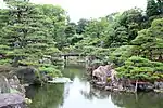 A pond with stones along the sides located in a garden with pine and other trees.