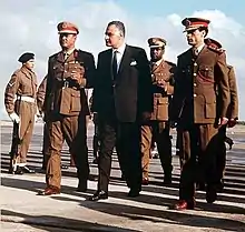 Three men walking side by side. The man in the middle is wearing a suit, while the two to his side are wearing military uniforms and hats. There are a few other men in uniform walking behind them