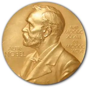 A golden medallion with an embossed image of a bearded man facing left in profile. To the left of the man is the text "ALFR•" then "NOBEL", and on the right, the text (smaller) "NAT•" then "MDCCCXXXIII" above, followed by (smaller) "OB•" then "MDCCCXCVI" below.