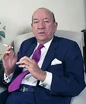 Noël Coward sitting part side on, but facing the camera; his hands are part-raised in gesticulation, and he smokes a cigarette.