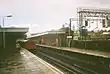 People walking on a wet railway platform with grey lampposts on either side of a railway track on which a red train is running all under a blue sky