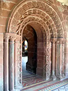 A highly decorated arched Norman doorway with a further, lower, arch beyond leading to a room with a window containing stained glass.