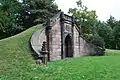 A small knoll rises from the left and his held back by a brown, gothic stone wall with gothic entrance. A shield is carved above the entrance and contains, among other words, the letters VAIL.