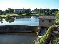 A body of water with a curved stone dam. At the right of the dam is a small stone house, underneath which water flows. There are buildings on land behind the dammed water.