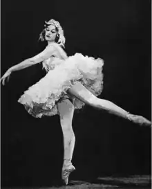 Photograph of a ballerina in a short tutu and feathered headdress, standing en pointe on her right leg with her left arm and left leg extended at 45-degree angles from the vertical line of her right leg and torso.