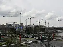 At least a dozen cranes rise over buildings under construction. In the foreground, cars are driving by on existing streets, and a city skyline rises in the background.