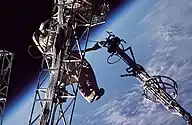 A man dressed in a spacesuit clings to a truss structure, manipulating a second truss with his left hand. Cabling can be seen running along both trusses, and the Earth's horizon can be seen in the background against the blackness of space.