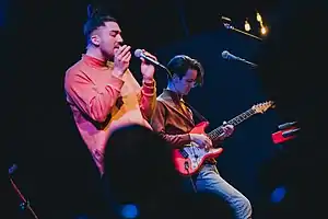 Two young men on stage, one singing into a handheld microphone and the other playing an electric guitar