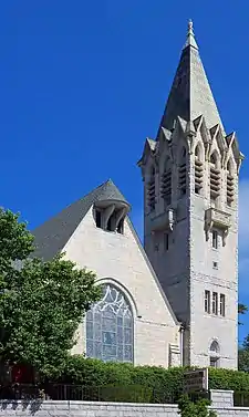 A bright-colored stone building with a steep pointed roof and a tall square pointed tower on its right. It has a large stained-glass window in front and some decorative touches.