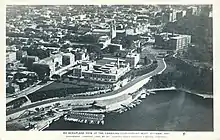 A road winds along the edge of a small cliff, with a mid-rise urban area behind it. Several prominent government buildings are located on both sides of the road. A bay of water is at the bottom of the cliff, where the Ottawa and Rideau Rivers meet.