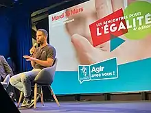 Ouissem is sitting on a chair on stage and speaking into a microphone; behind him is the poster of the event "Rencontres de l'Égalité" ; He is wearing a black t-shirt and blue jeans