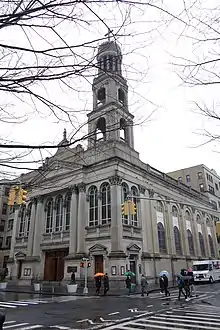 Three quarter view of a church at the corner of two streets. A bell tower rises atop the corner of the church