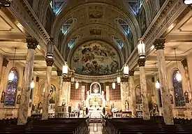 Nave of a church, looking toward the altar. A large mural above the altar is framed by columns. Pews are in the foreground.