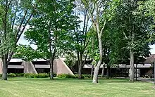 A long green lawn stands before a low brick building, mostly obscured by trees.