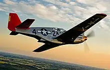 A single nosed-propeller plane is in mid flight above land. The red nose faces right and the underside of the plane is slightly in view. The plane has black propellers, grey base paint, and black letters reading A42. The 4 and 2 are separated by a United States roundel in black with a central white star. The roundel is also visible on the tops of the planes wings. The full length of the right side of the plane is visible.