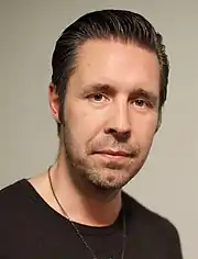 A dark-haired man wearing a black shirt poses for the camera in a blank background