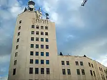 Ground-level view of a 10-story building; the exterior has a tan hue with dark windows and a higher front side