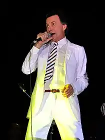 A man in a white tuxedo is facing left holding a microphone