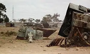 Pieces of a destroyed tank, notably the gun turret, lie on a sandy landscape.