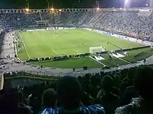 A football match taking place in a packed stadium with an athletics track, viewed from a stand behind one of the goals; it's night and the ground is floodlit.