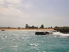  Picture of a sandy shore delineated from both sides by dark brown rocky formations. The sandy shore is at the edge of a wavy blue colored body of water. At the top the light blue sky shows some white clouds and some sea birds.