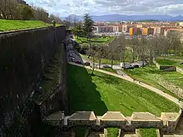 Photo shows an old wall at left and modern buildings at right with mountains in the distance.