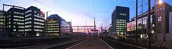 Railroad tracks surrounded by lit office buildings as the sun sets below the horizon