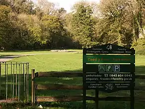 Cairn in the distance in sunshine, with trees in leaf to its left, right and rear. To its front lies flat ground of short grass. An asphalt path leads from the left past the cromlech. The shaded foreground has a kissing gate, a wooden fence and a Forestry Commission welcome sign in Welsh (first) and English.