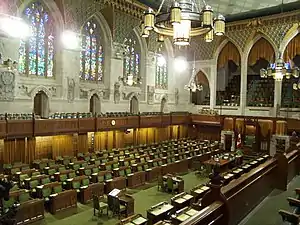 A view of a building interior with a chandelier hanging from the ceiling, four stain-glass windows on the left wall, and a large number of brown desks and green chairs on the floor.