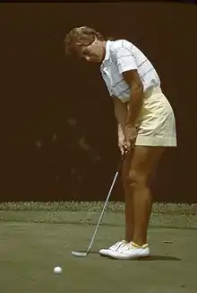 A blonde-haired woman in a mostly white shirt and yellow shorts is hitting a putt with a putter with white golfing shoes on