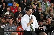 A middle-aged man in a white shirt and black and yellow striped tie speaks into a microphone on stage in front of a crowd. (from Wisconsin)