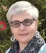 headshot of a middle aged white woman with short grey hair and somewhat fancy glasses standing in front of some greenery