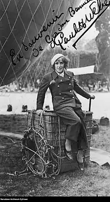 Young woman sitting on the edge of a hot air balloon basket that is set on the ground. She's wearing pants, a double-breasted jacket and a beret. There is a part of a hot air balloon looming behind her.